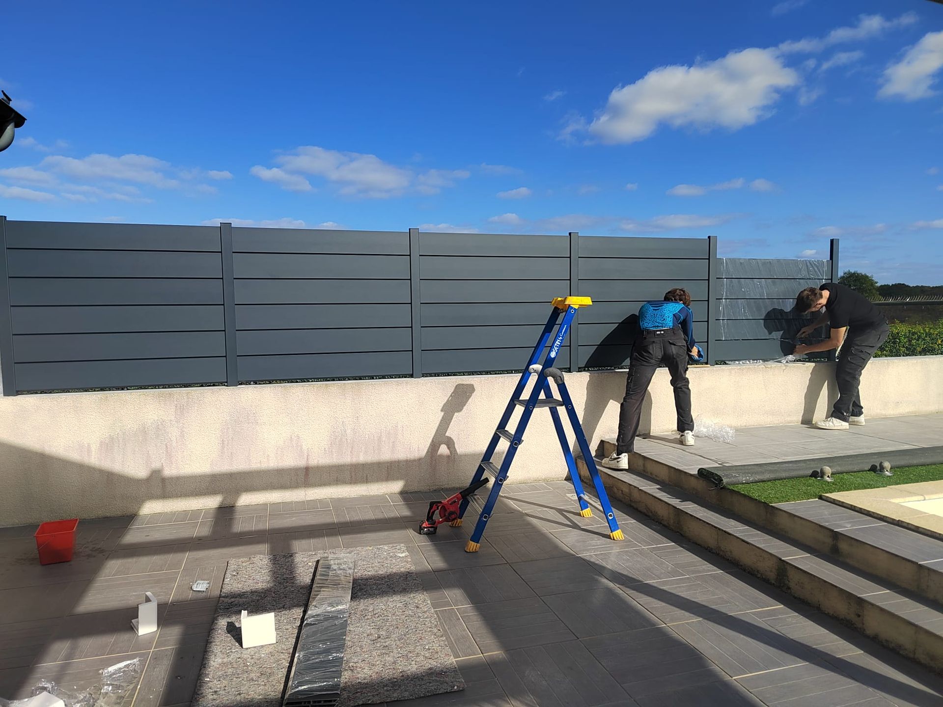 Deux personnes installent une clôture grise à lattes horizontales sur un mur en béton.
