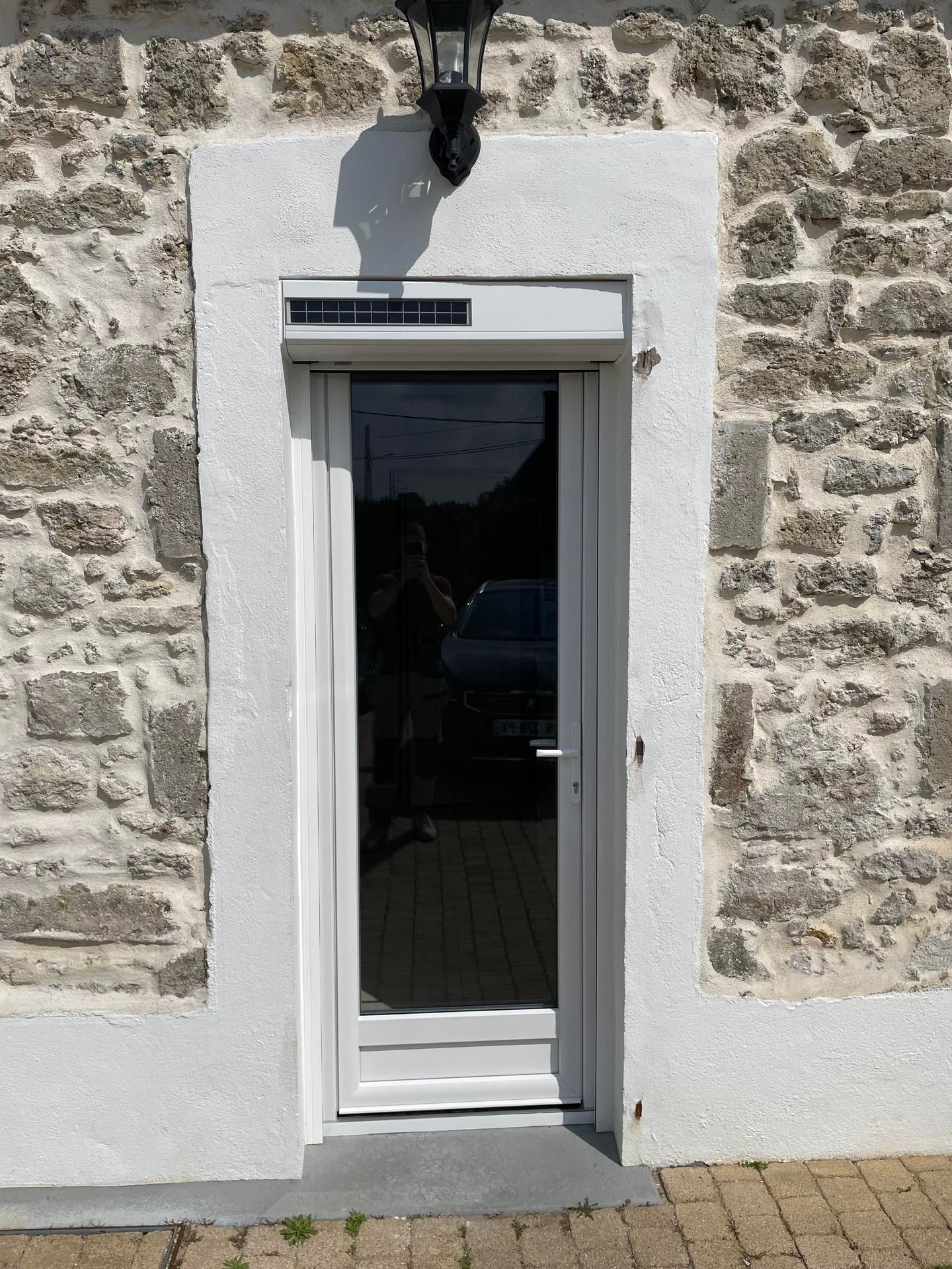Porte vitrée à cadre blanc avec store intégré, encastrée dans un mur en pierre. Un luminaire noir est installé au-dessus de la porte.