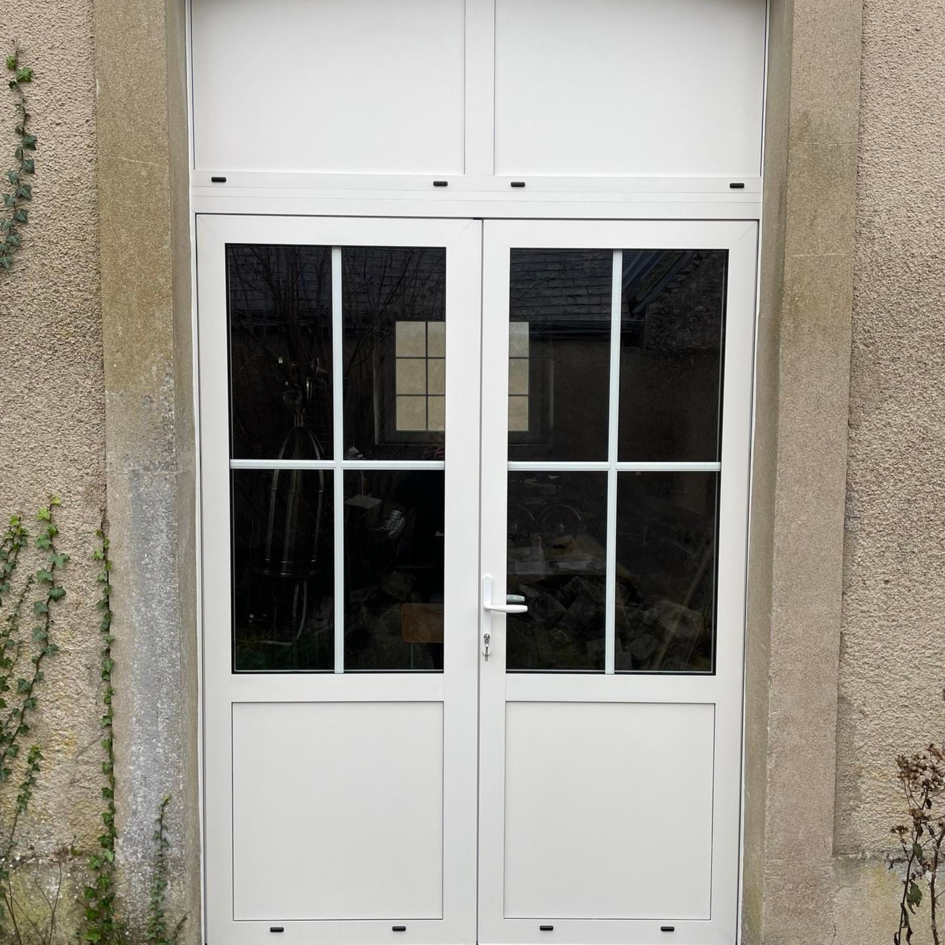 Portes-fenêtres blanches à vitrage et imposte, encastrées dans un mur en pierre.