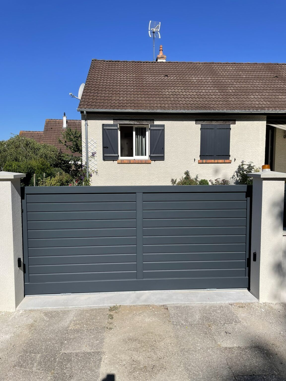 Portail d'allée à lattes horizontales grises devant une maison de couleur claire avec un toit marron.