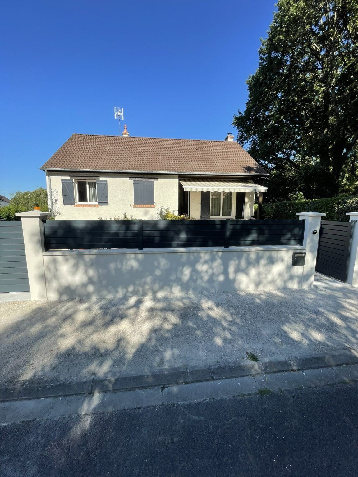 Maison au toit marron, aux volets gris et à la clôture noire, par une journée ensoleillée.