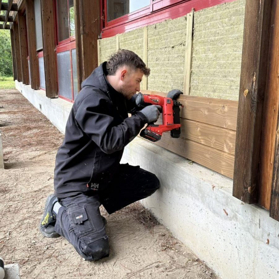 Une personne installant un bardage en bois sur l'extérieur d'un bâtiment, à l'aide d'un outil électrique, tout en étant à genoux.