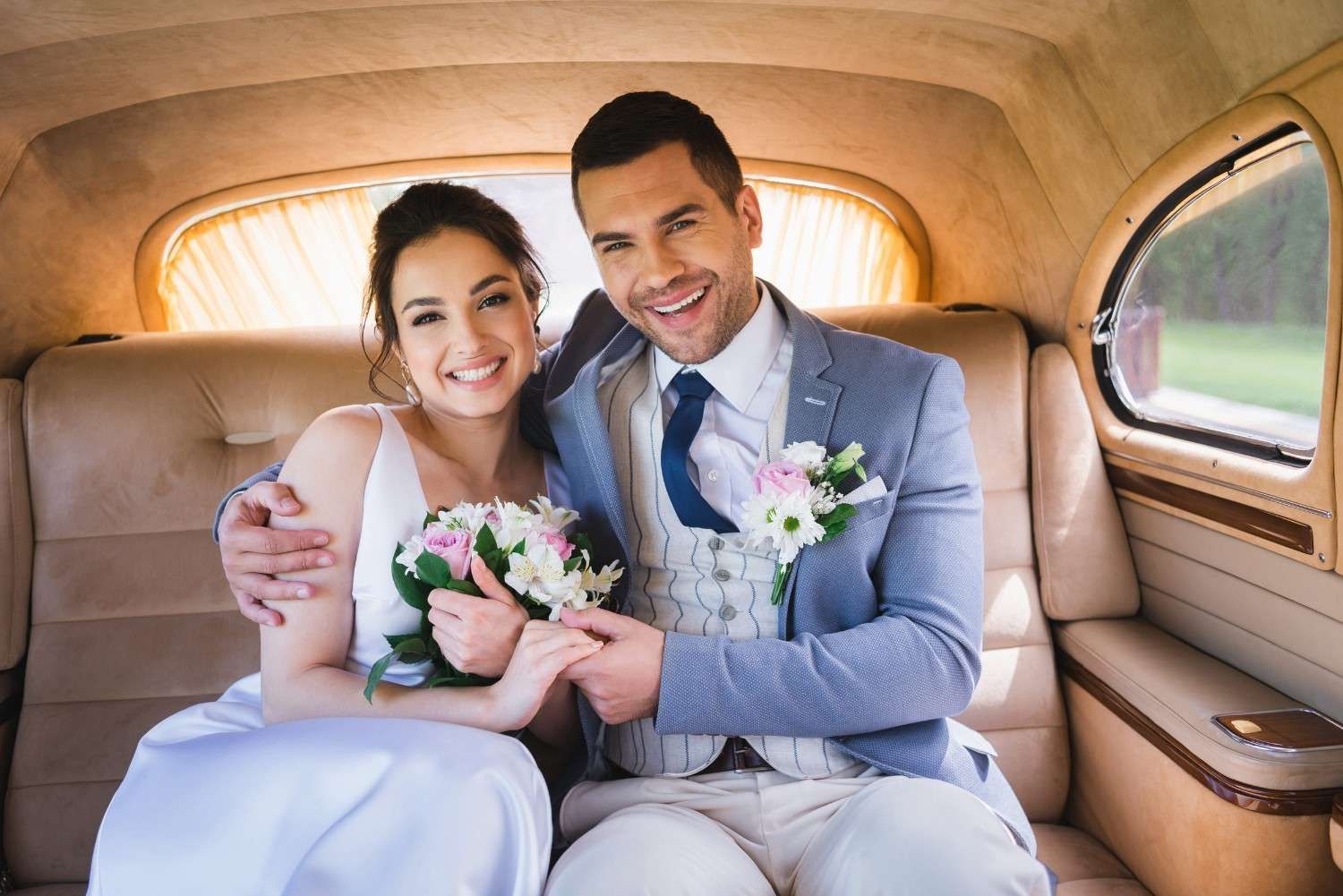 Una pareja sonriente, vestida de novios, está sentada dentro de un coche antiguo, sosteniendo un ramo de flores.