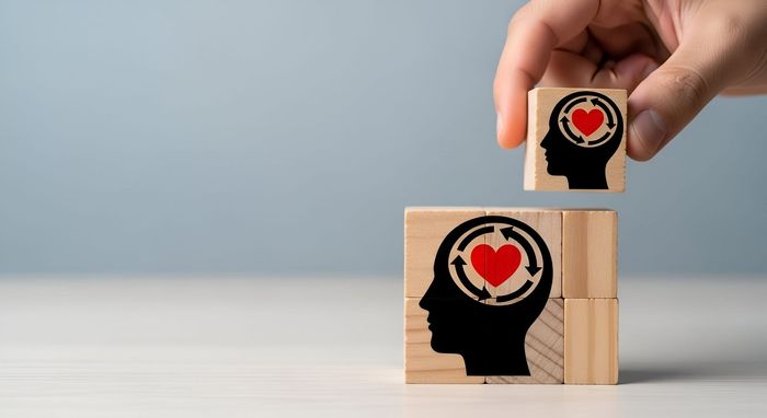 Una mano coloca un bloque de madera con un icono de cabeza y corazón sobre una torre de bloques a juego