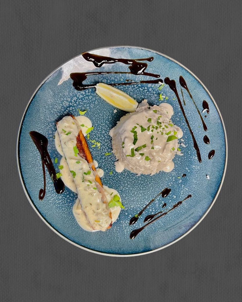 Assiette bleue avec du poisson, un quartier de citron et un filet de sauce brune sur une table grise.