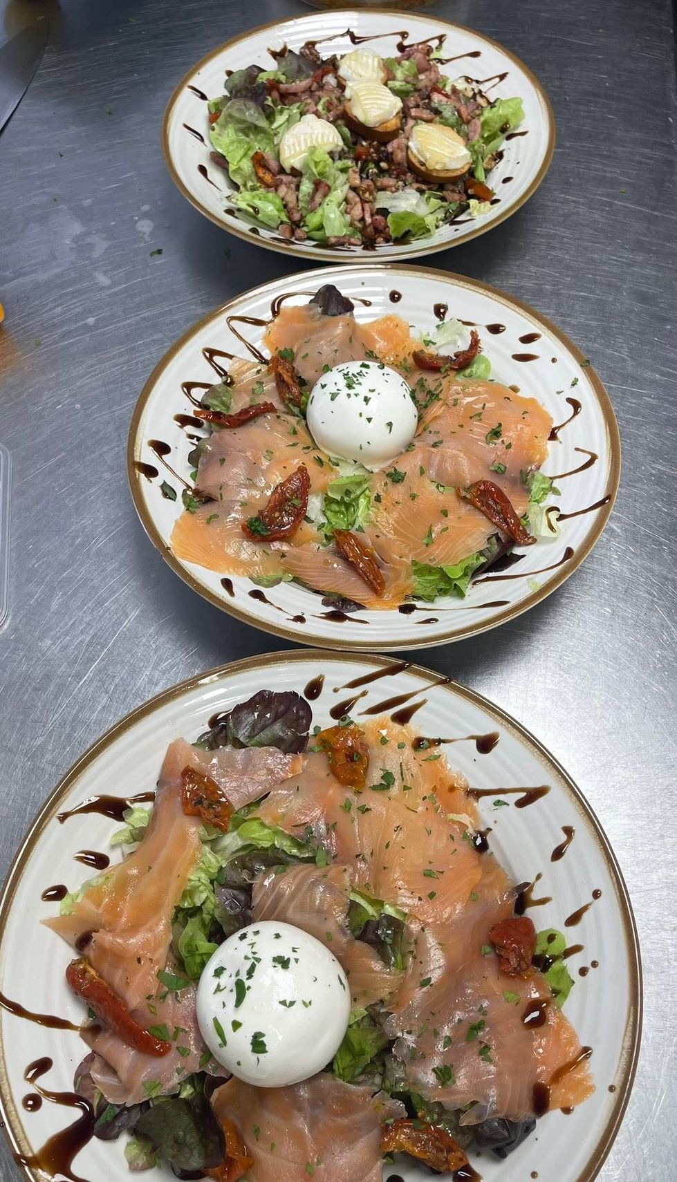Deux assiettes de saumon avec sauce crémeuse, légumes verts et œufs pochés, et une assiette de salade chèvre chaud, toutes présentées sur une table en métal.
