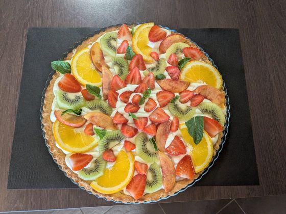 Tarte aux fruits garnie de fraises, de tranches d'orange, de kiwi et de crème, présentée sur un plat foncé.