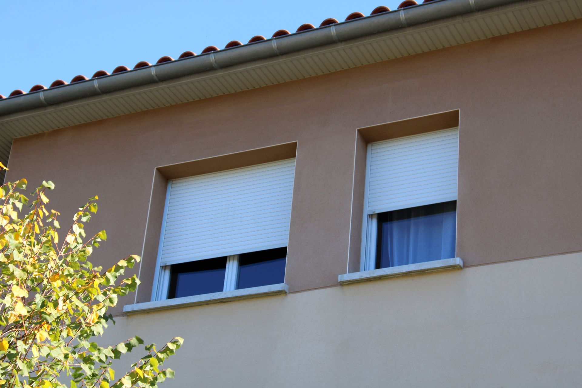 Deux fenêtres fermées sur le mur d'une maison beige avec un toit en tuiles, avec une branche d'arbre feuillue au premier plan.