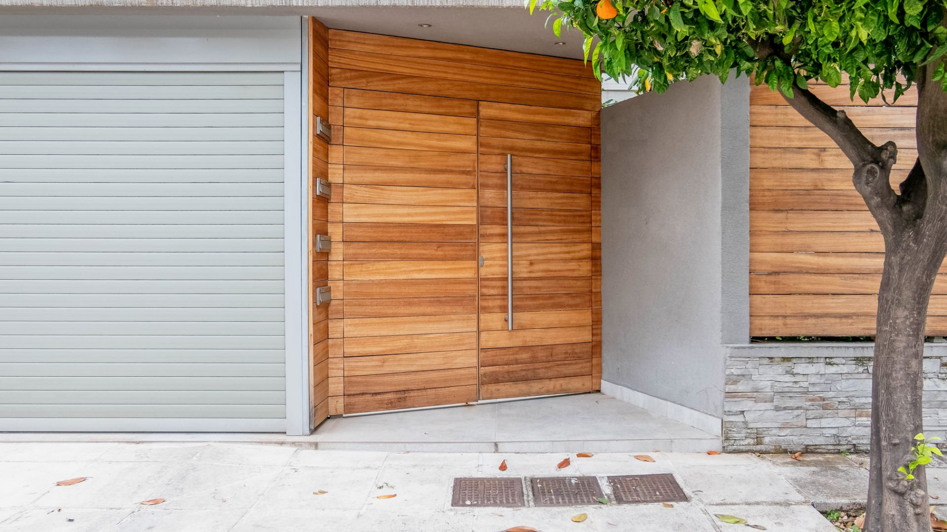 Entrée de maison moderne avec porte d'entrée en bois, porte de garage grise et arbre près d'un mur en pierre.