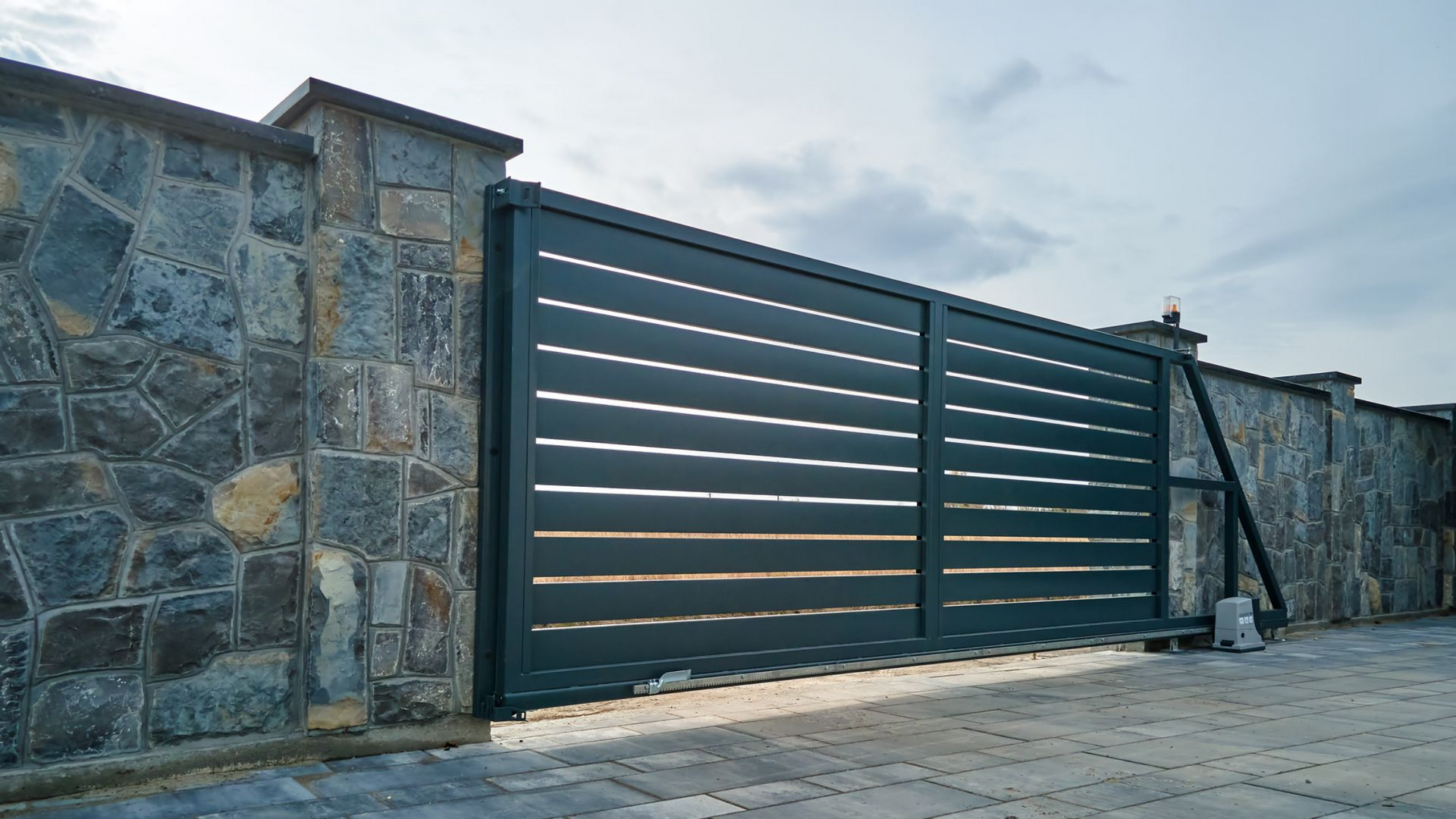 Portail coulissant en métal bleu foncé avec un mur en pierre le long d'une allée pavée au crépuscule.