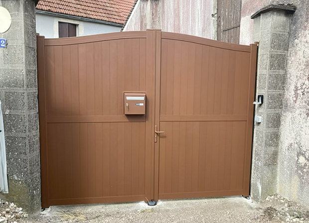 Portail double en métal marron avec une boîte aux lettres installée entre des murs en pierre dans une ruelle étroite