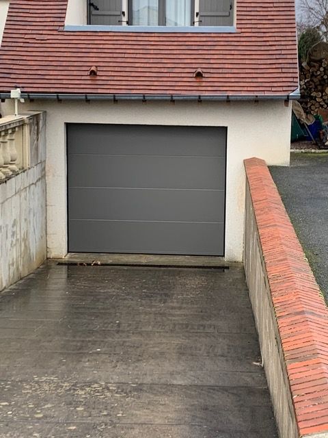 Porte de garage grise fermée sur une maison blanche avec une allée pavée et de petits arbustes.