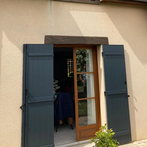 Des volets bleus ouverts encadrent une porte en bois sur une maison beige, avec de la verdure à l'entrée.