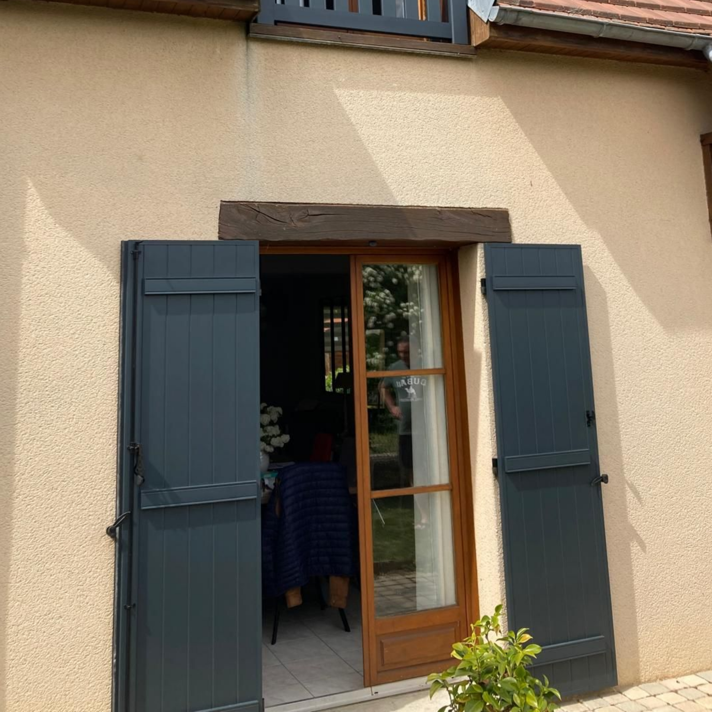 Fenêtre en bois avec volets bleu foncé sur un mur en stuc beige, partiellement ouverte sur une vue du jardin