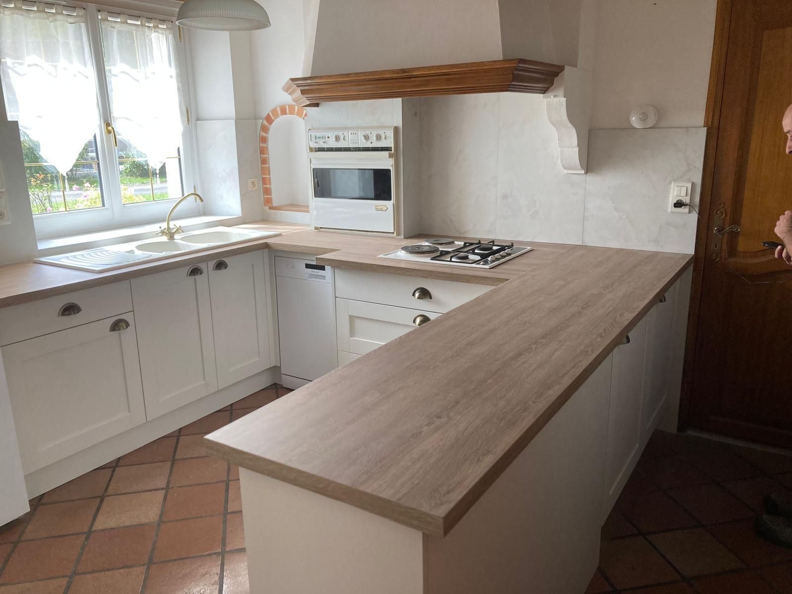Cuisine lumineuse avec placards blancs, carrelage en terre cuite au sol, îlot central avec plan de travail en bois, fenêtre, four et cuisinière à gaz.