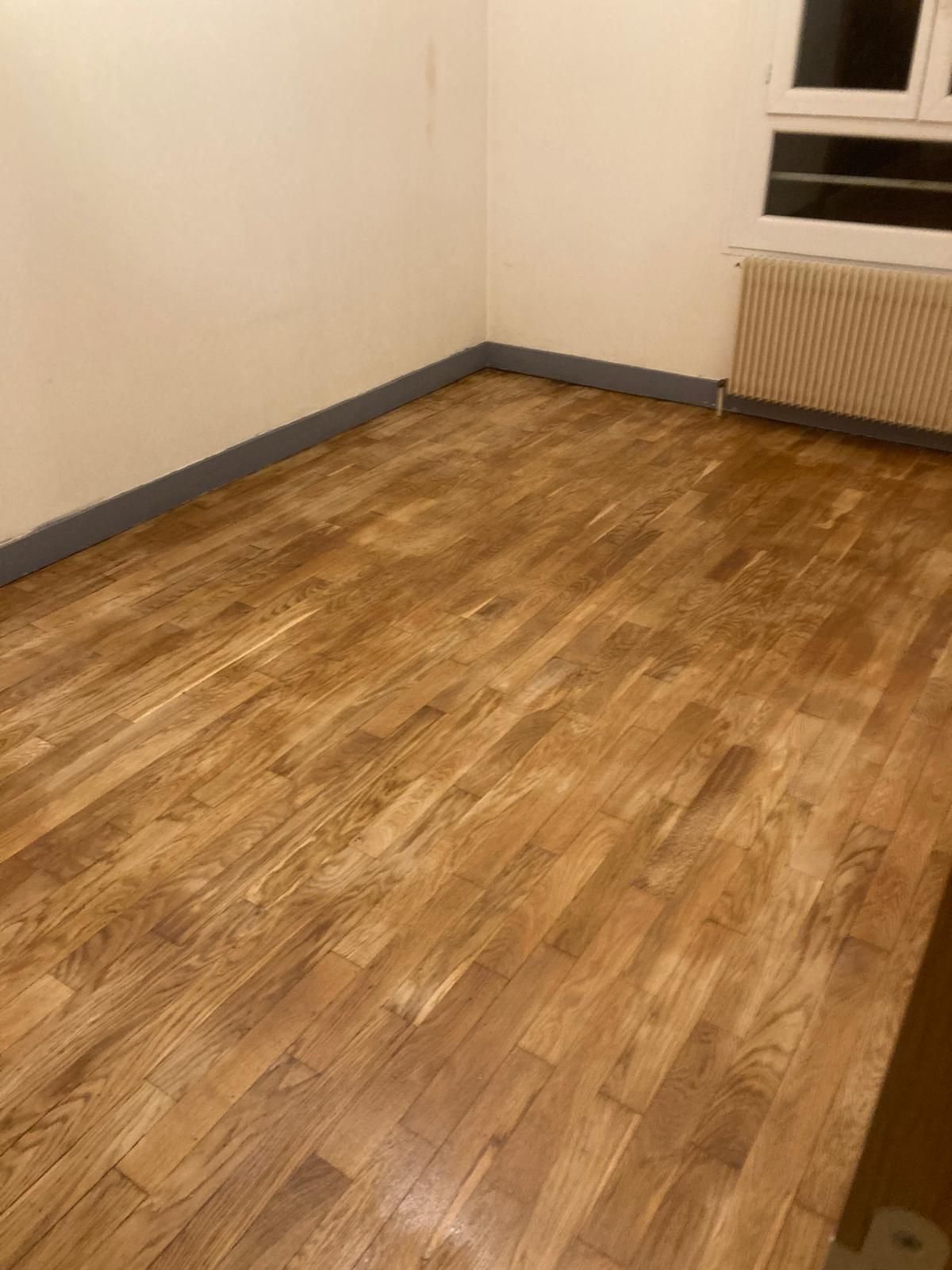 Pièce vide avec parquet, murs beiges et une fenêtre au-dessus d'un radiateur.
