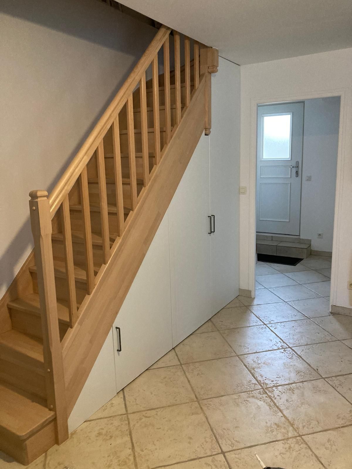 Un escalier en bois se trouve à côté d'une porte blanche fermée, dans un couloir carrelé menant à une petite pièce.