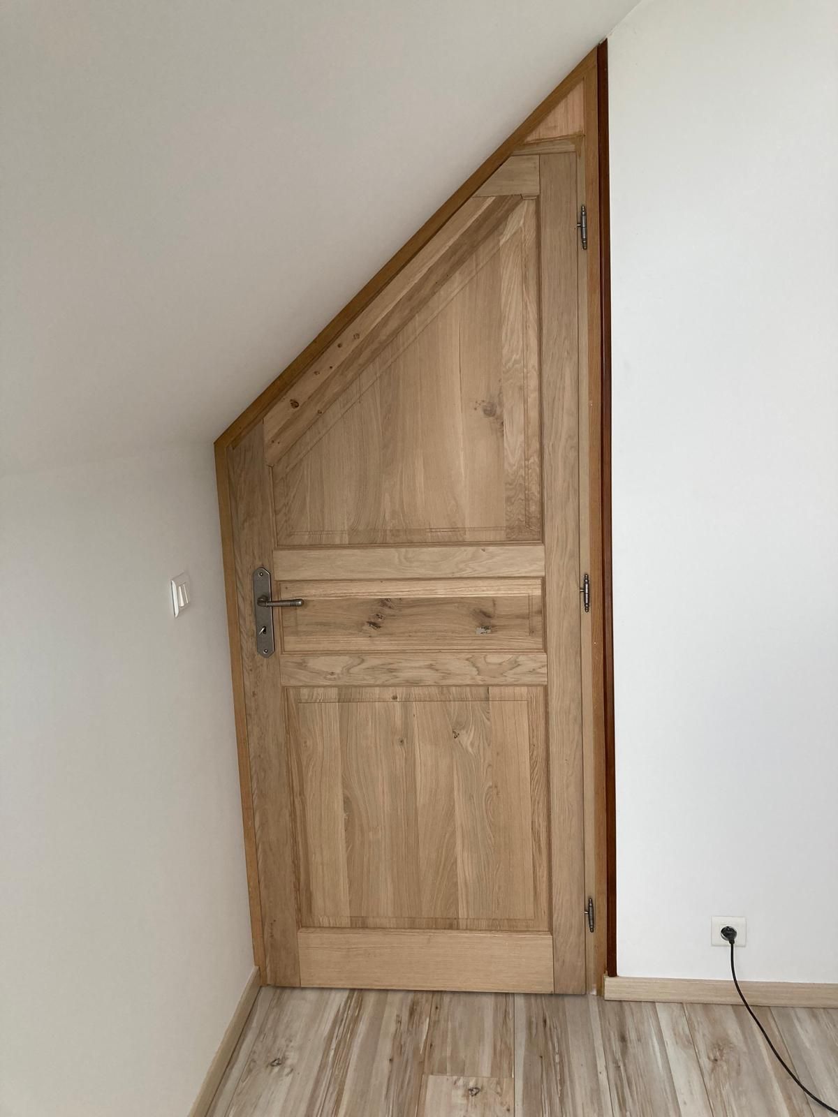 Porte en bois sous un plafond mansardé dans une petite pièce blanche au sol carrelé