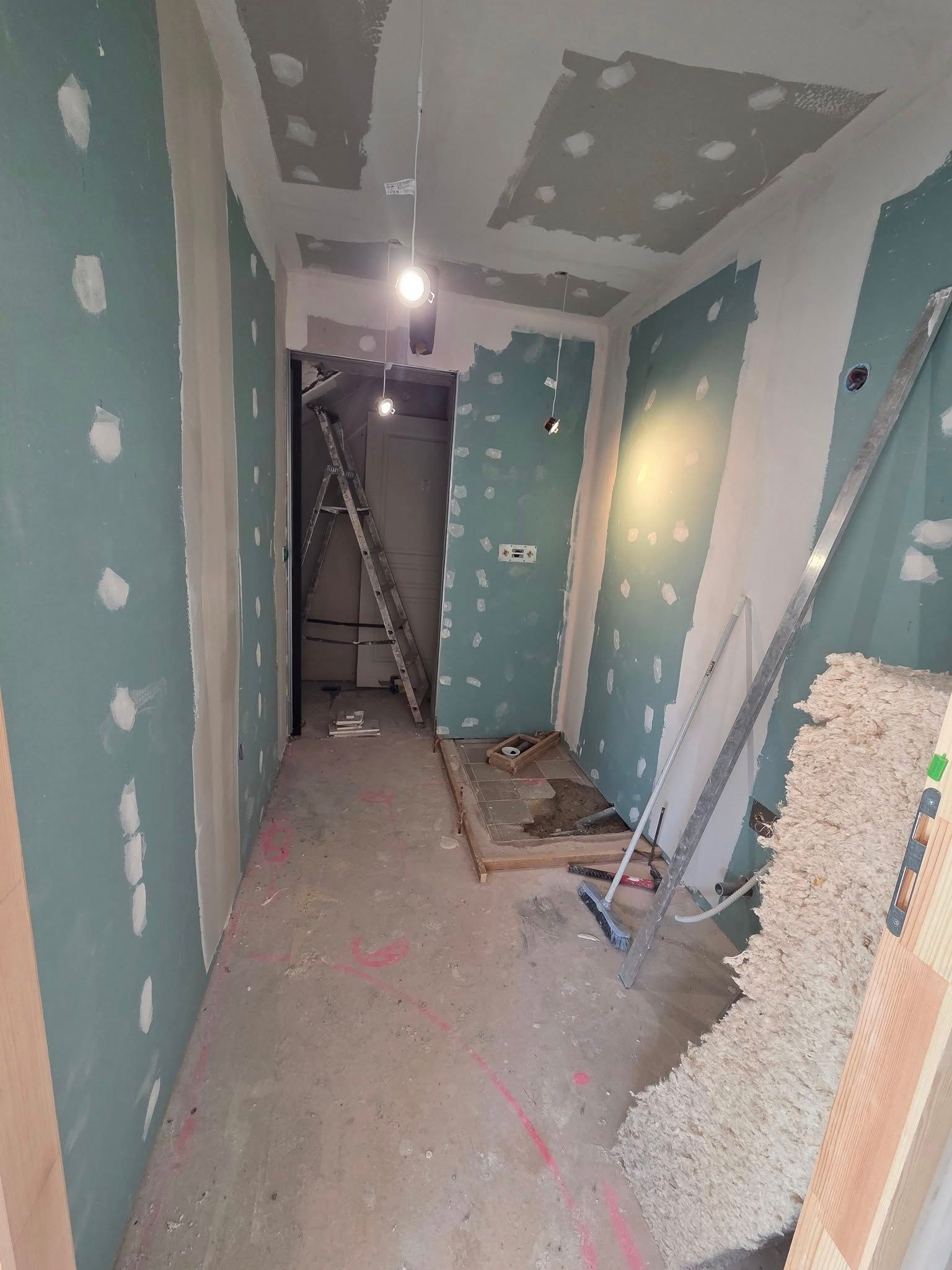 Couloir en construction : cloisons sèches vertes, taches blanches, échelle, outils et une porte au bout.