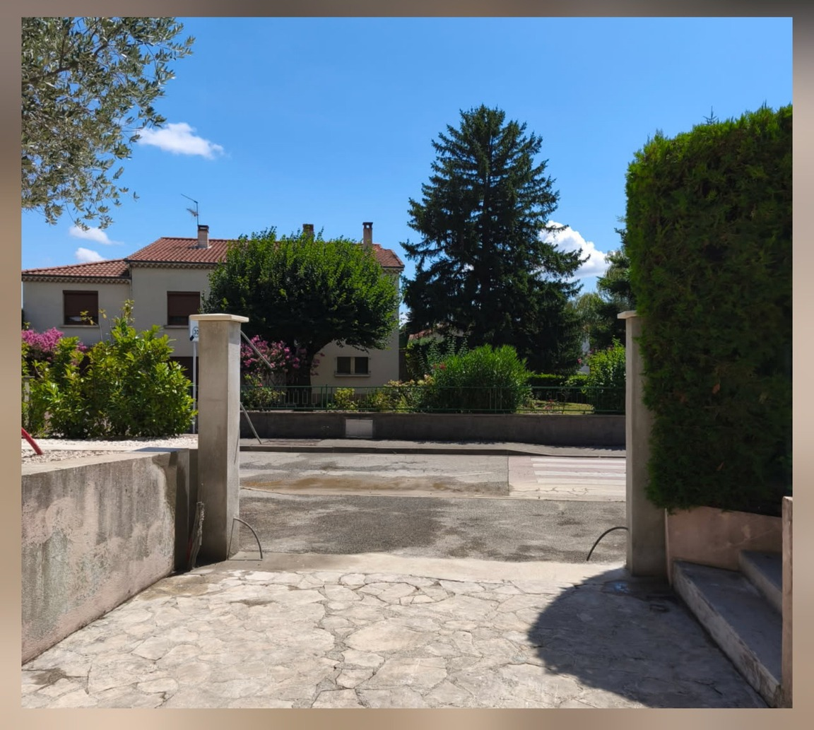 Vue depuis une allée, en direction d'une rue résidentielle. Une maison et des arbres sont visibles sous un ciel bleu.