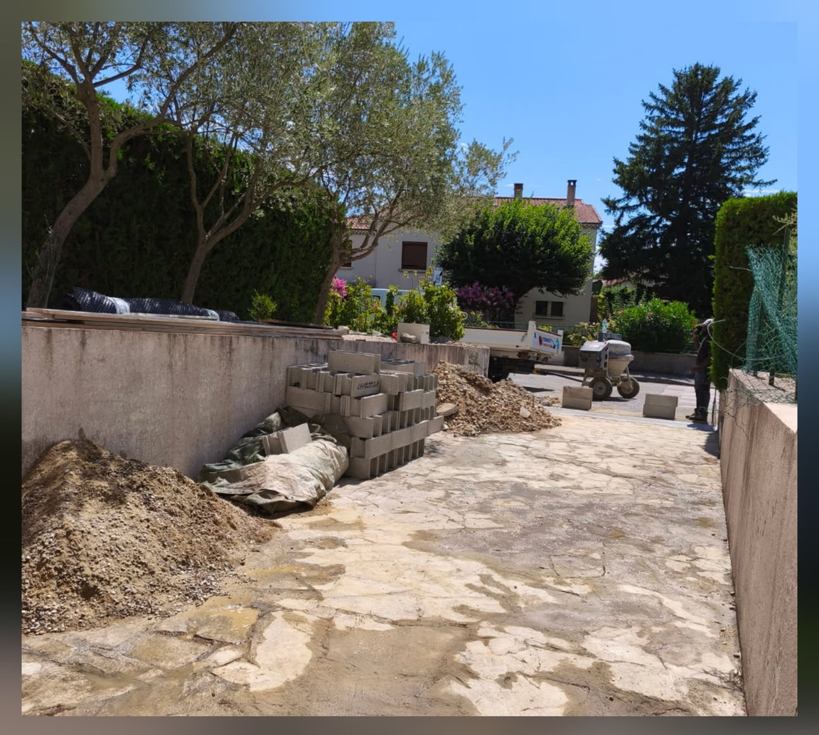Arrière-cour en construction avec des blocs de béton, un tas de terre et une maison en arrière-plan par une journée ensoleillée.