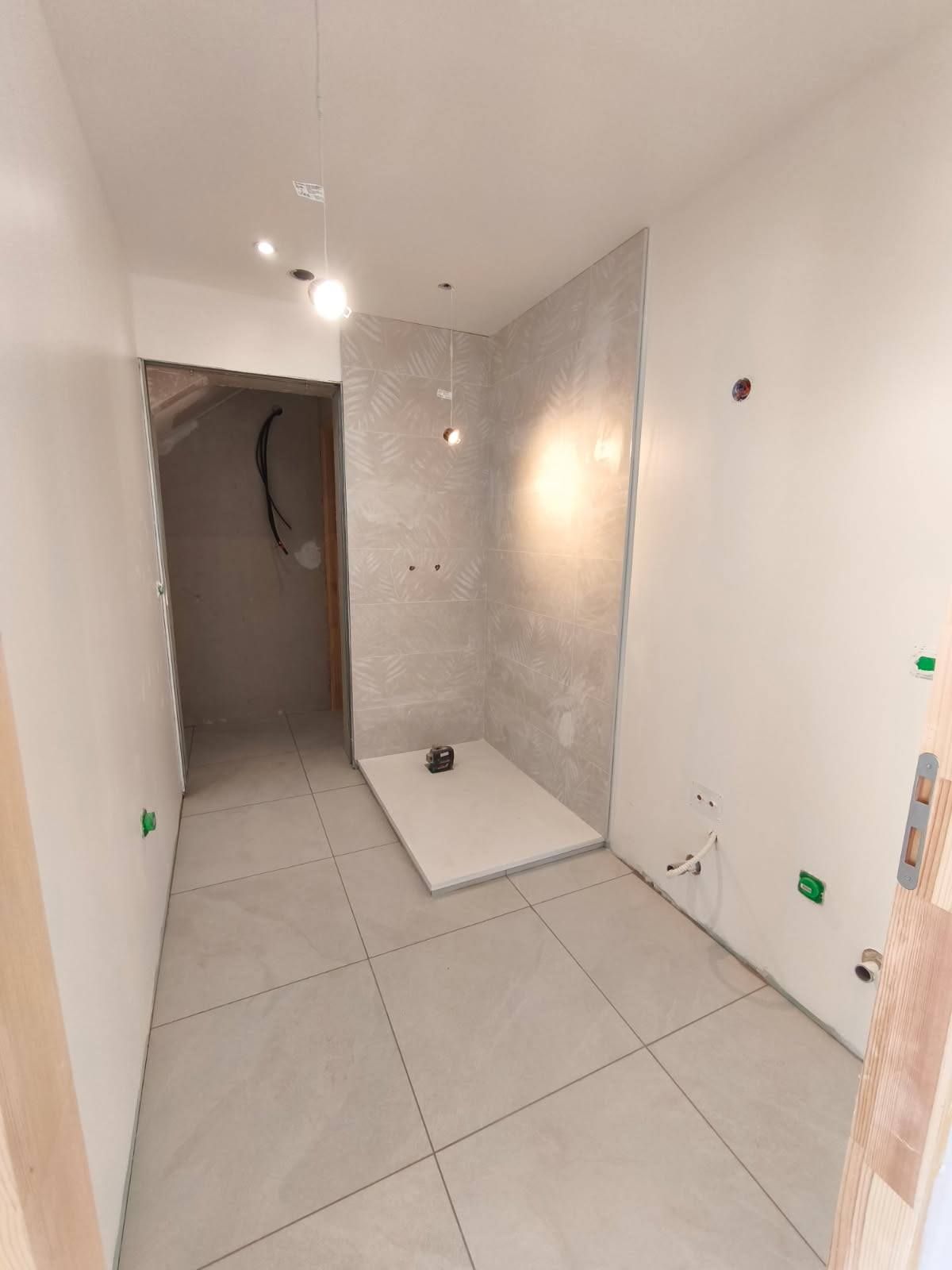 Salle de bains en construction, avec carrelage gris, receveur de douche et plomberie apparente sur les murs.