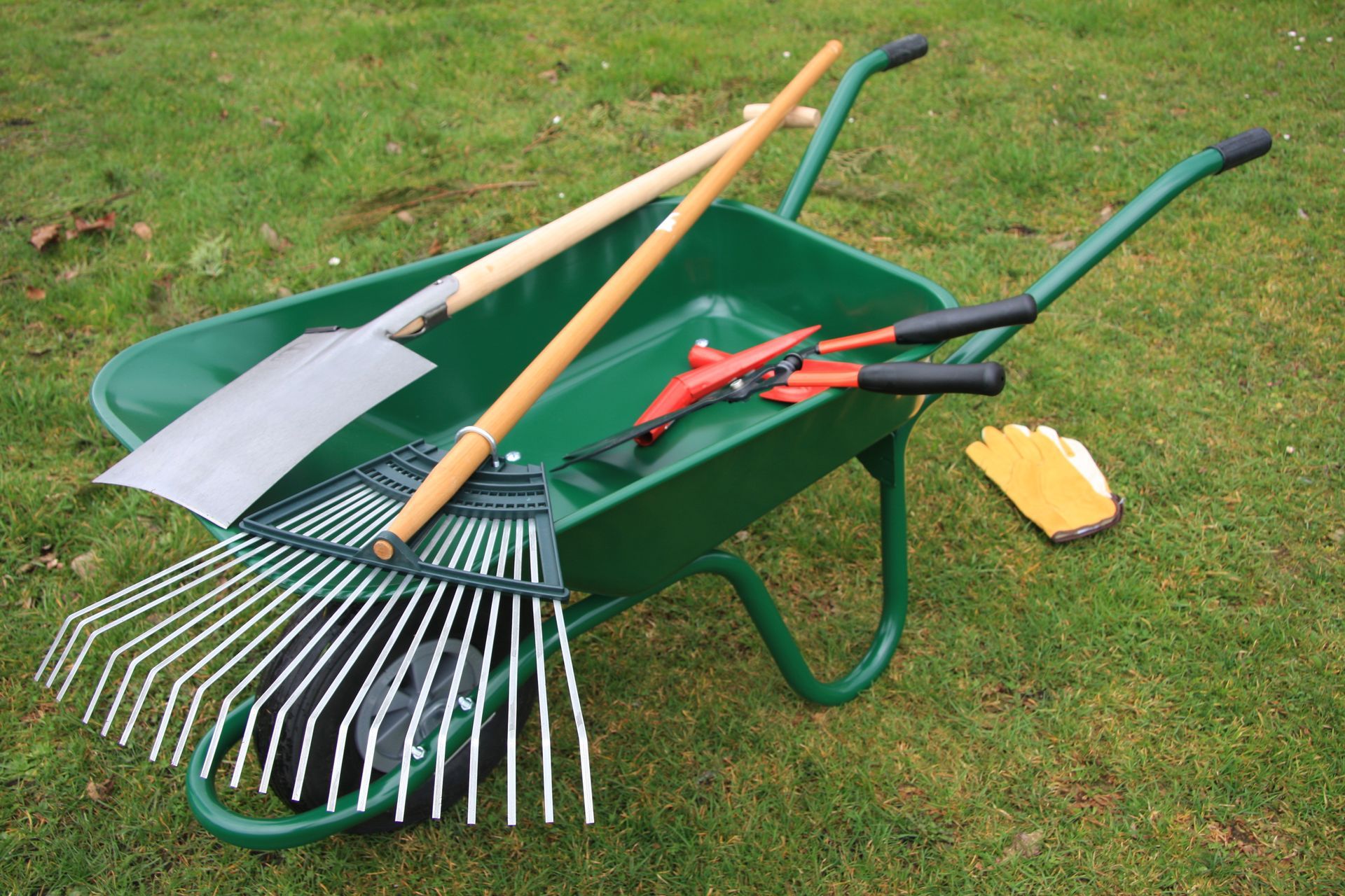 Outils de jardinage dans une brouette verte sur l’herbe : une pelle, un râteau, un coupe-branches et des gants.