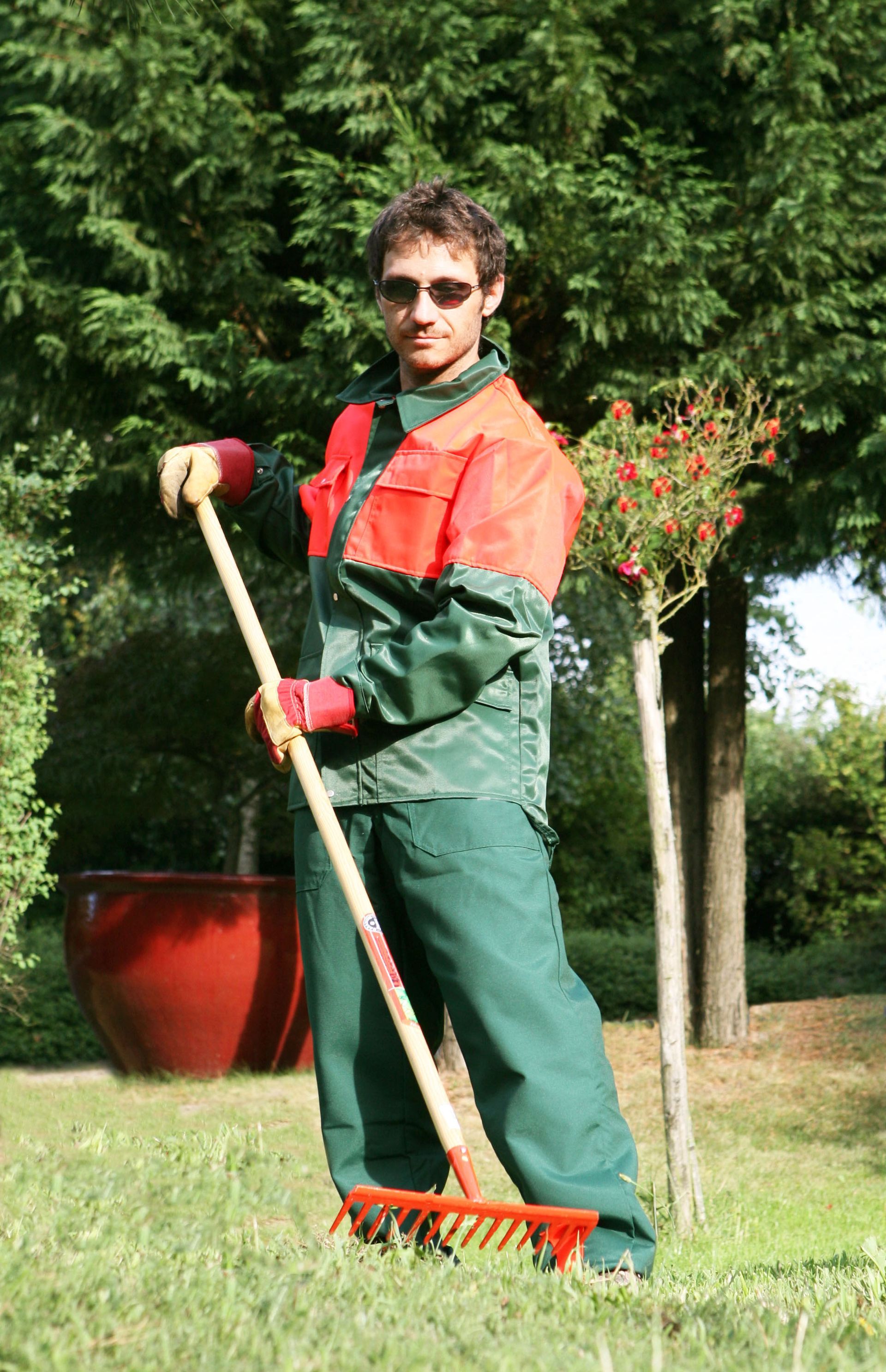 Personne en tenue de travail verte tenant un râteau dans un jardin herbeux avec des plantes en pot et des arbres.