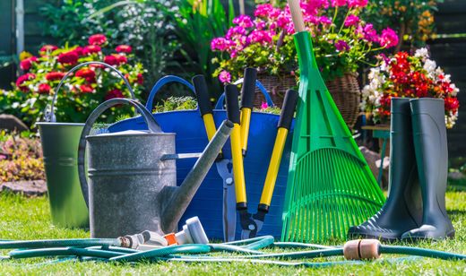 Des outils de jardinage et des arrosoirs disposés sur l’herbe, avec des fleurs colorées en arrière-plan.