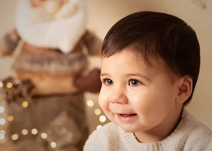 Niño pequeño sonriente con un suéter claro; decoración de Papá Noel y luces de hadas al fondo.
