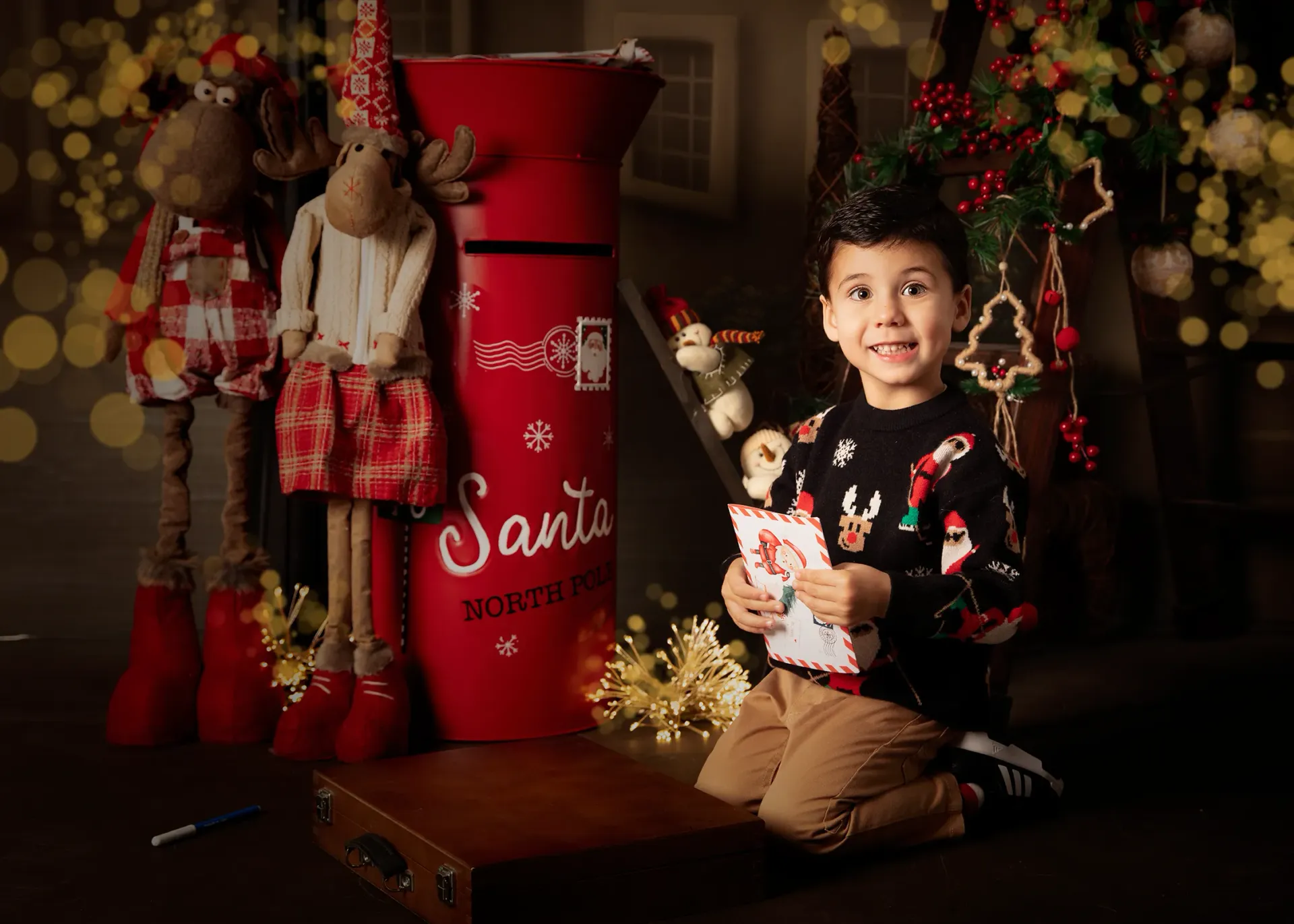 Niño arrodillado sosteniendo una carta, junto a un buzón rojo de Papá Noel, con decoración navideña de fondo.