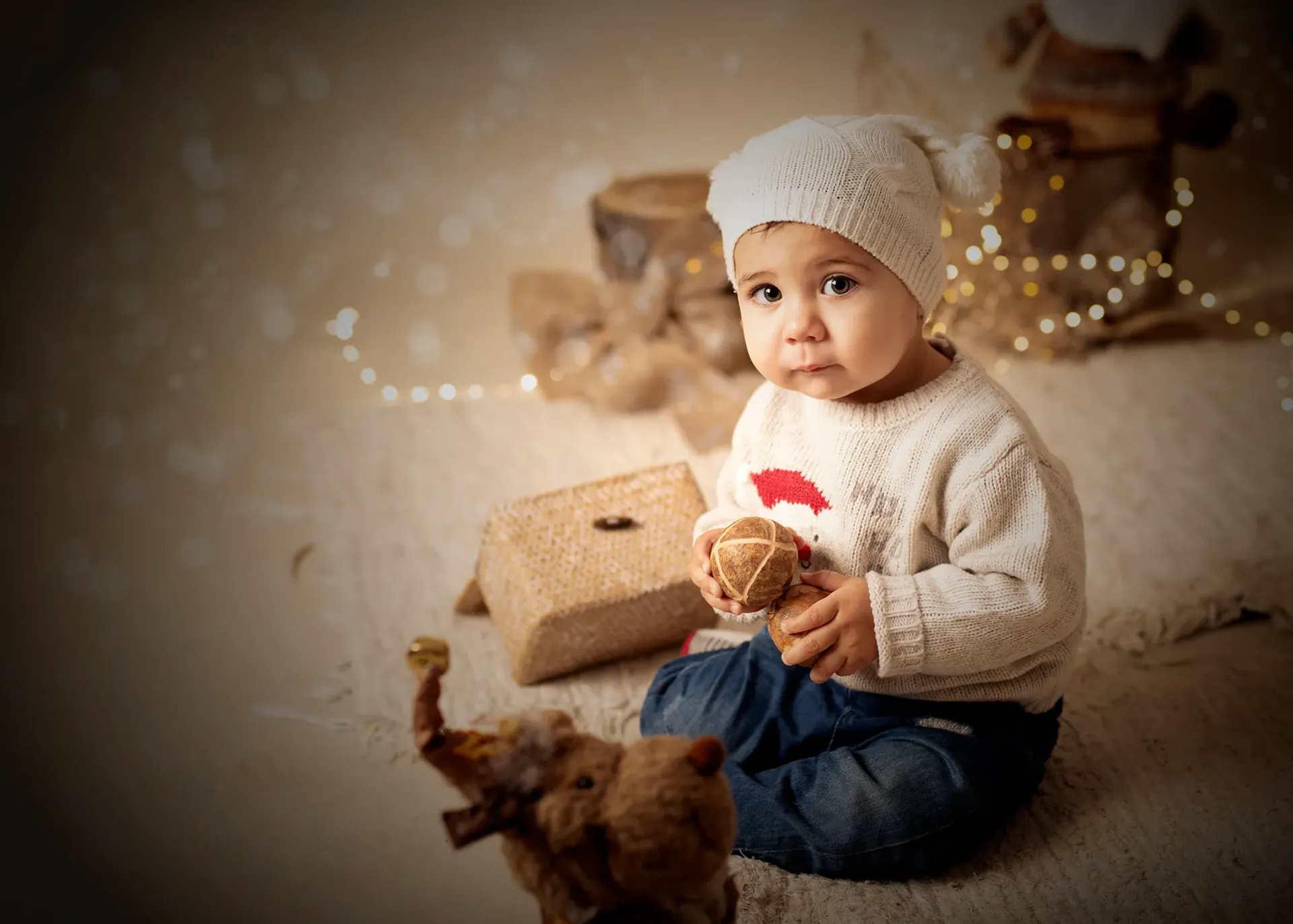 Un niño con gorro y suéter de lana está sentado, sosteniendo un objeto, cerca de un elefante de peluche y luces decorativas.