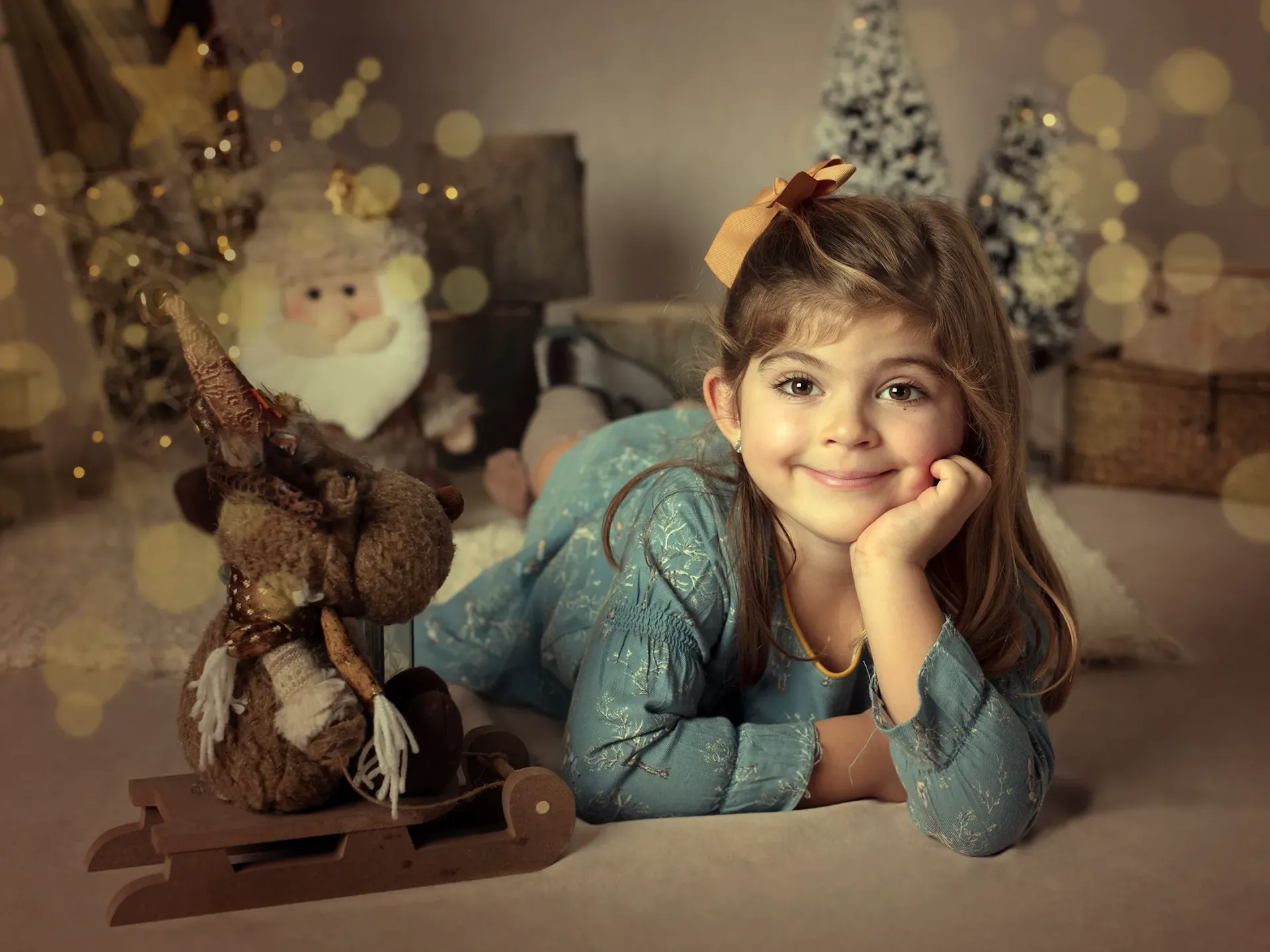 Una niña pequeña sonríe, recostada en una escena navideña con renos y Papá Noel.