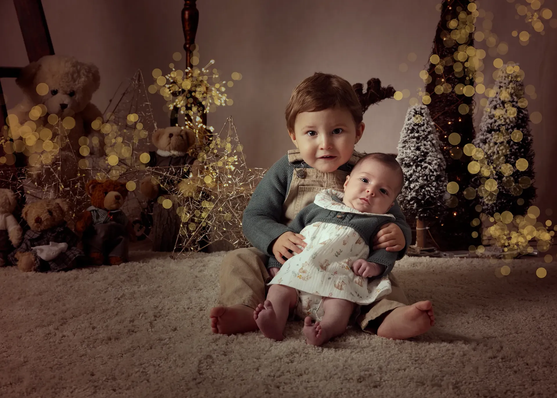 Niño con cuernos de reno sosteniendo a un bebé; ambos sonriendo, en un ambiente navideño con ositos de peluche y luces.