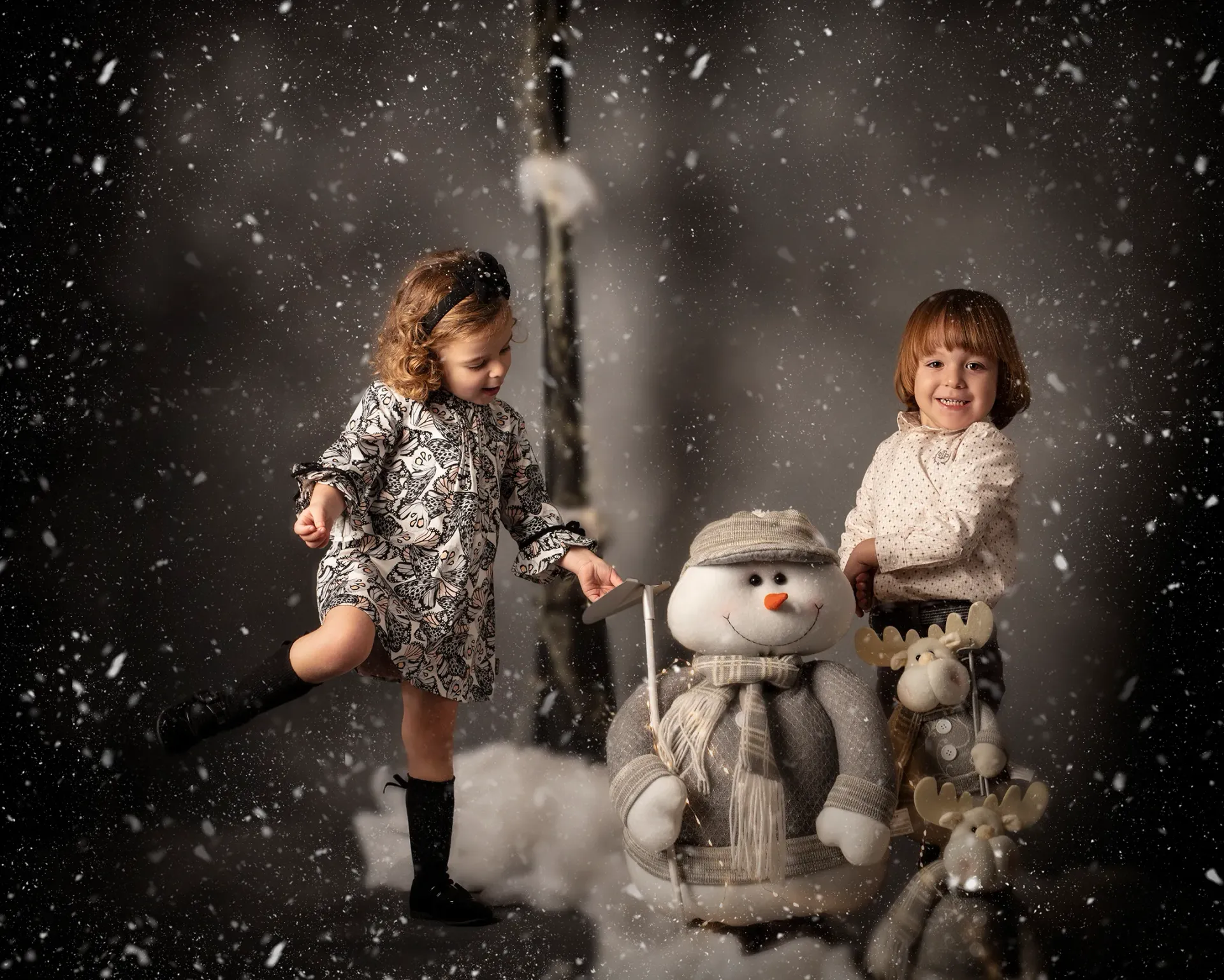 Dos niños con ropa de invierno posan con un muñeco de nieve; caen copos de nieve.