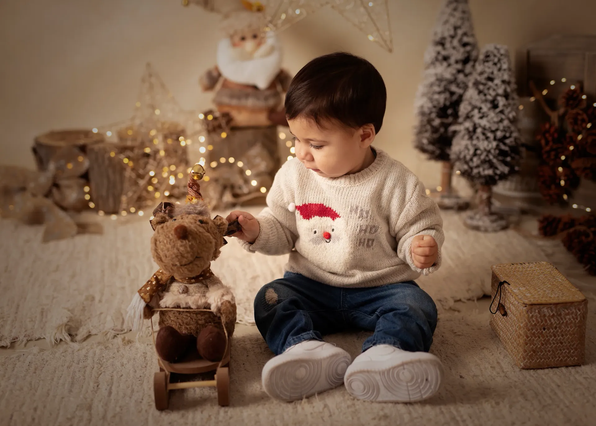 Un niño está sentado cerca de un juguete de reno entre adornos navideños, mirando hacia abajo.