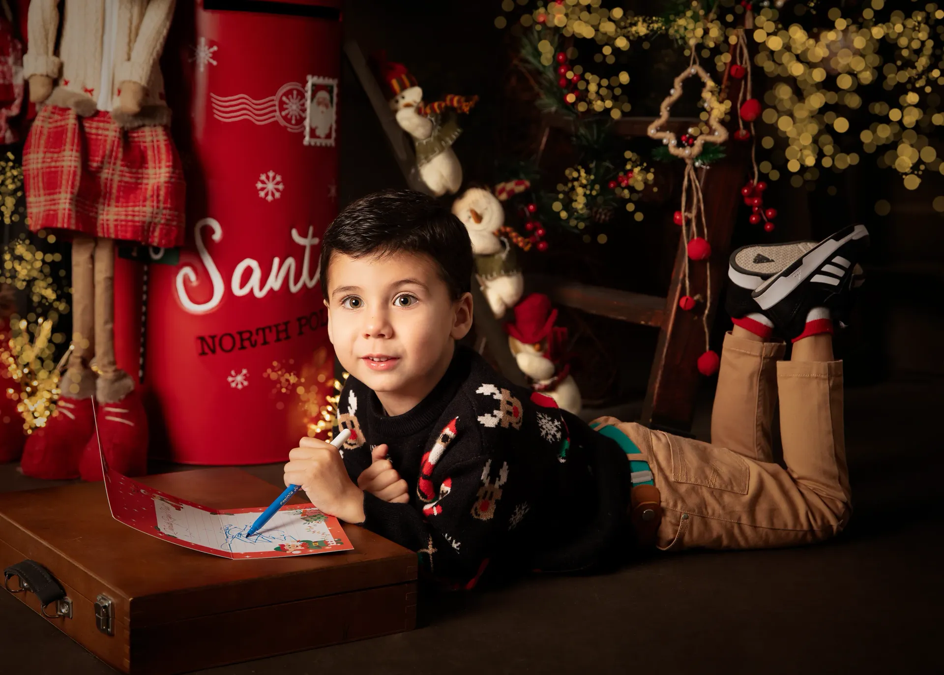 Niño escribiendo una carta en el suelo junto a un buzón rojo de Papá Noel y un árbol de Navidad.