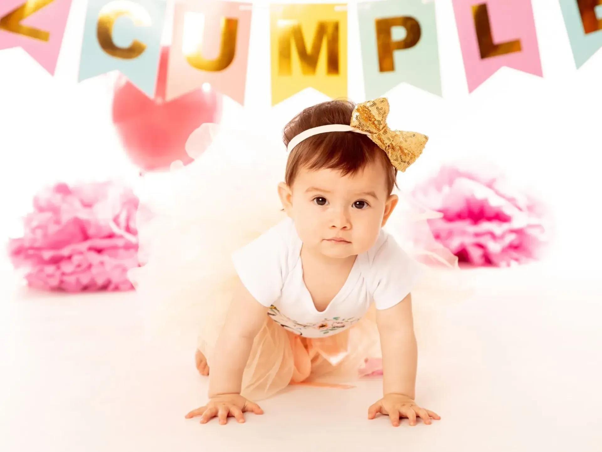 Bebé gateando, con diadema de lazo dorado y tutú, pancarta de cumpleaños al fondo.