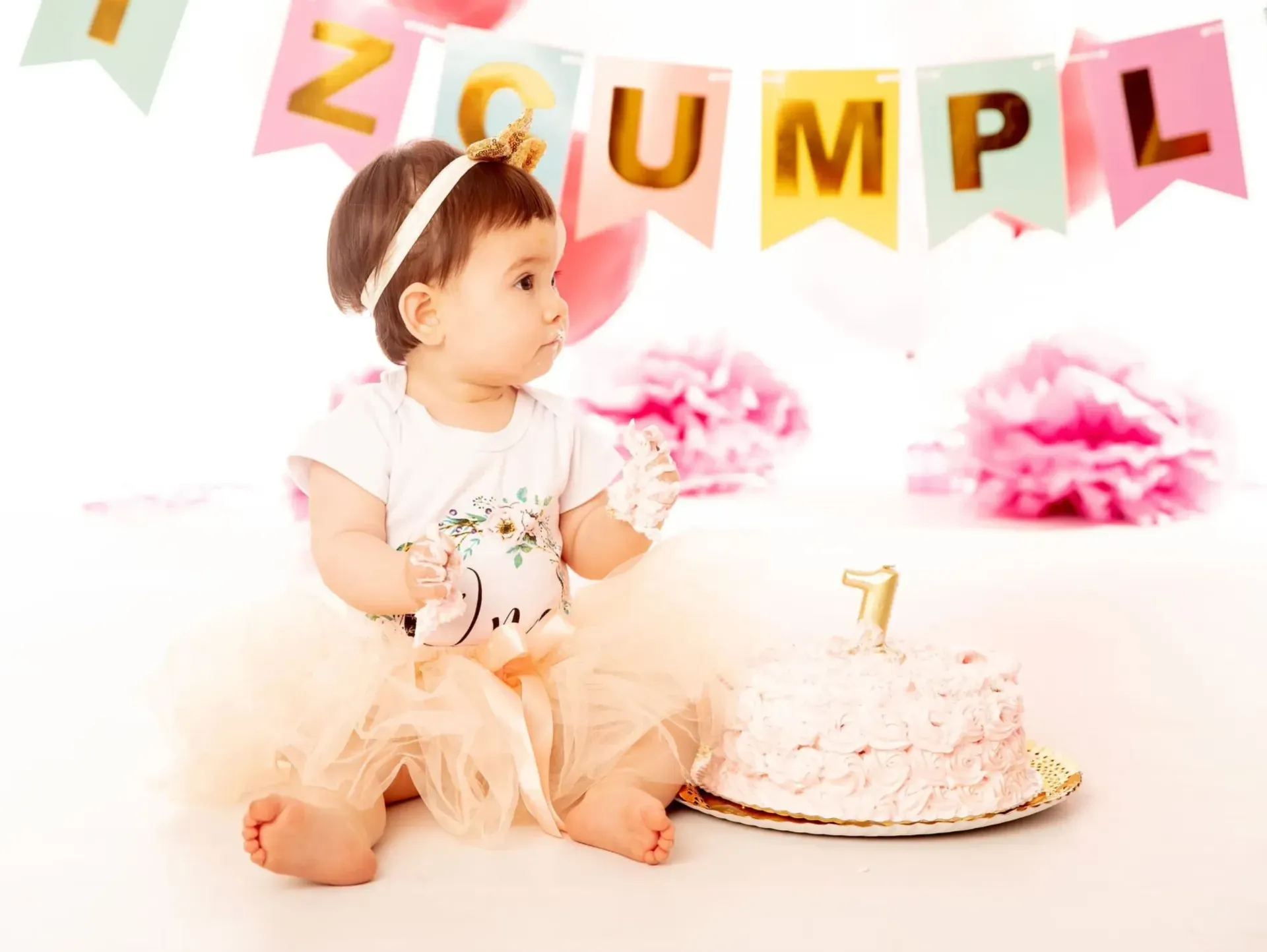 Una bebé está sentada cerca de un pastel, mirando algo fuera de cámara. Lleva una corona, un tutú y una pancarta que dice 