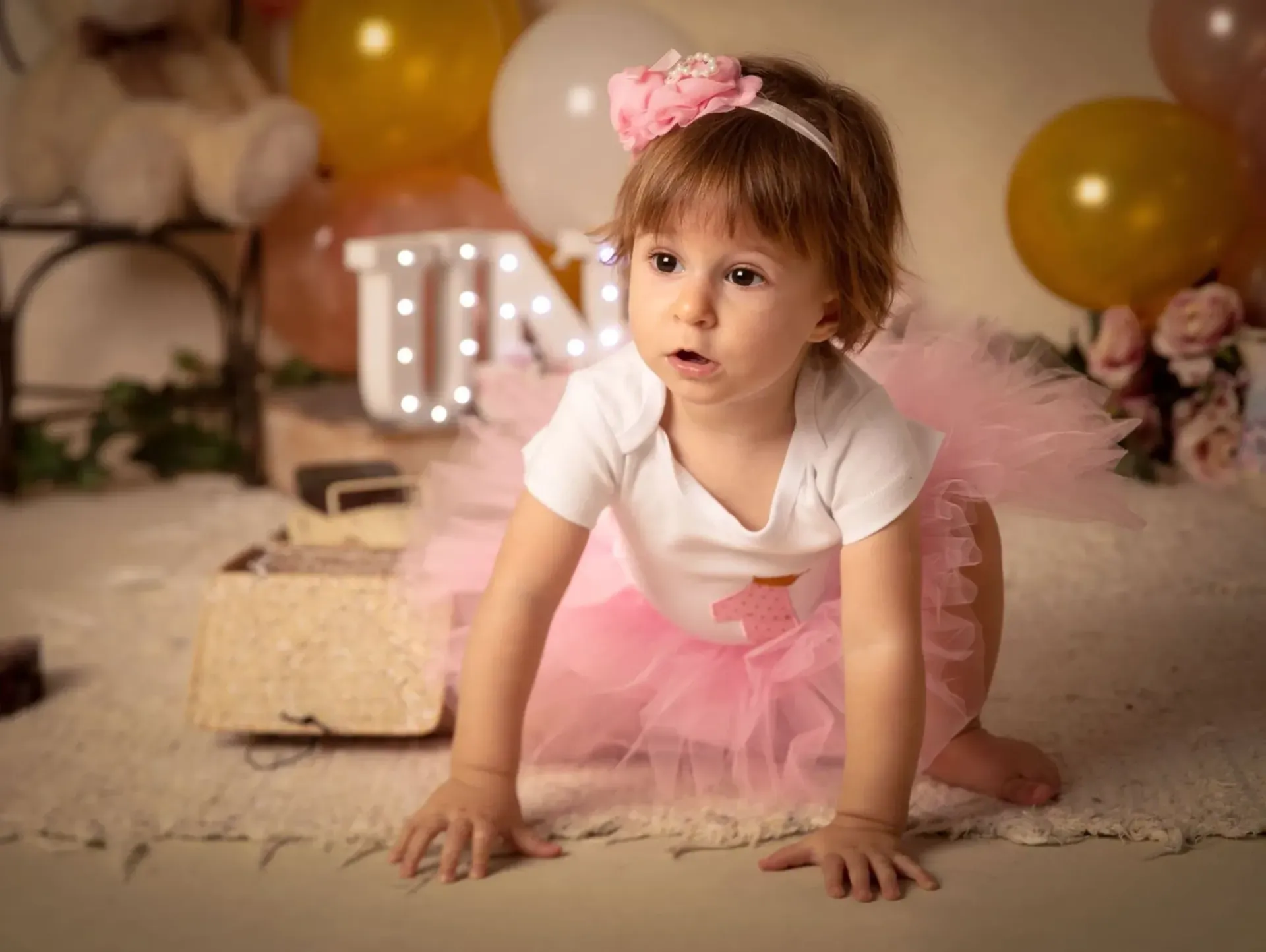 Niña gateando, vestida con tutú rosa, diadema y mameluco, rodeada de globos y un cartel que dice 