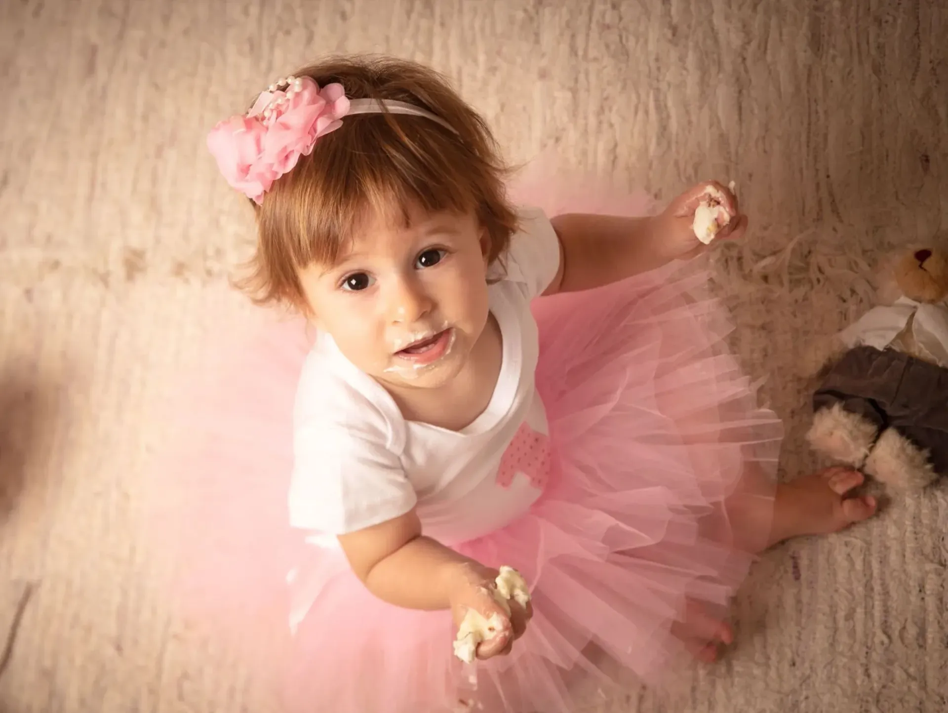 Niña pequeña con pastel en la cara, vestida con tutú rosa y diadema, mirando hacia arriba.