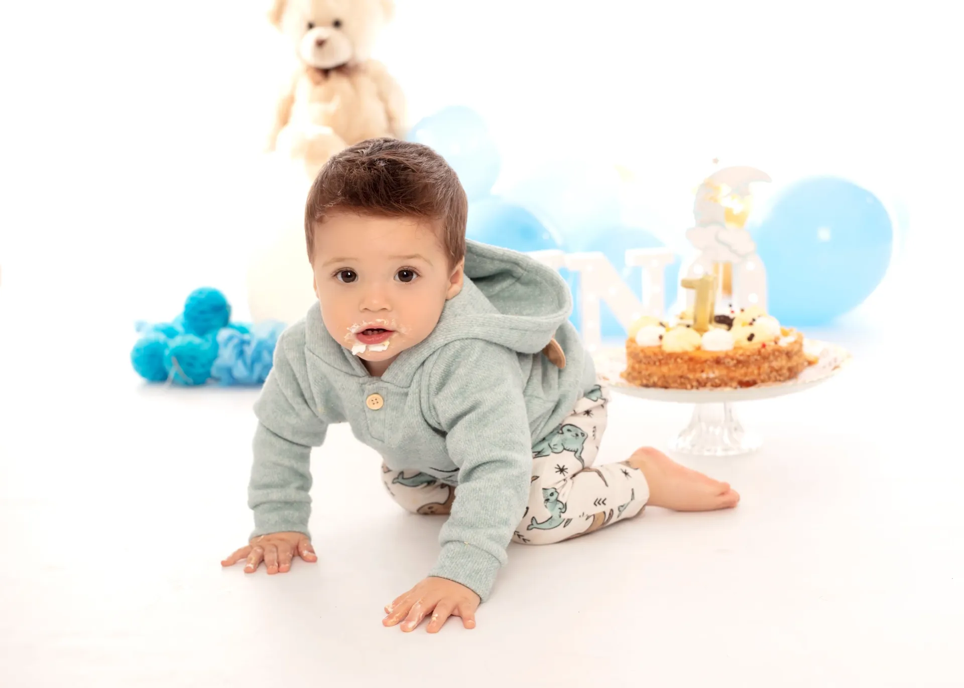 Bebé con glaseado en la cara gatea hacia la cámara, pastel de cumpleaños con el número “1” cerca, rodeado de globos azules.