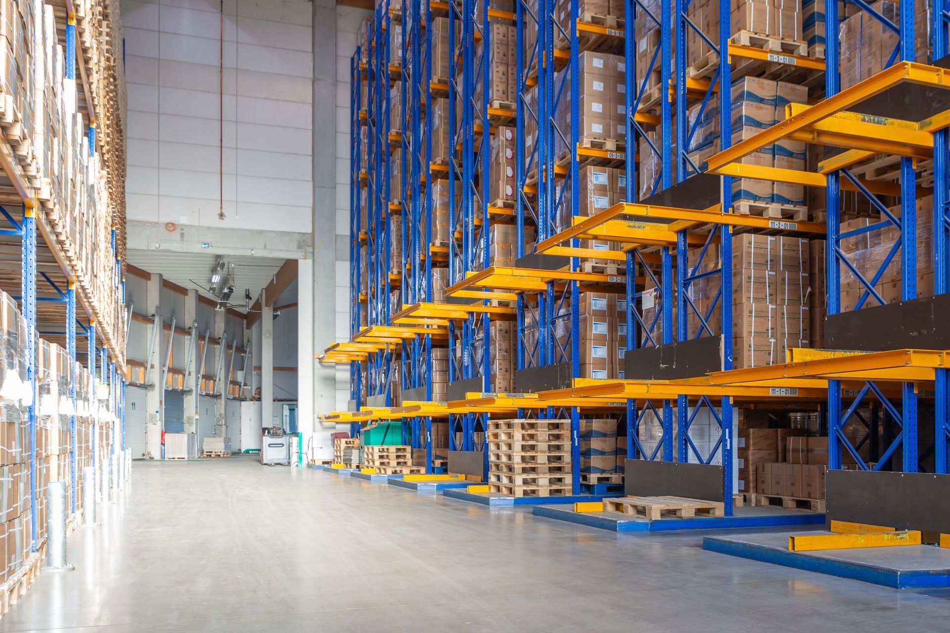 Intérieur d'entrepôt avec de hautes étagères bleues et jaunes supportant des palettes de marchandises empilées.