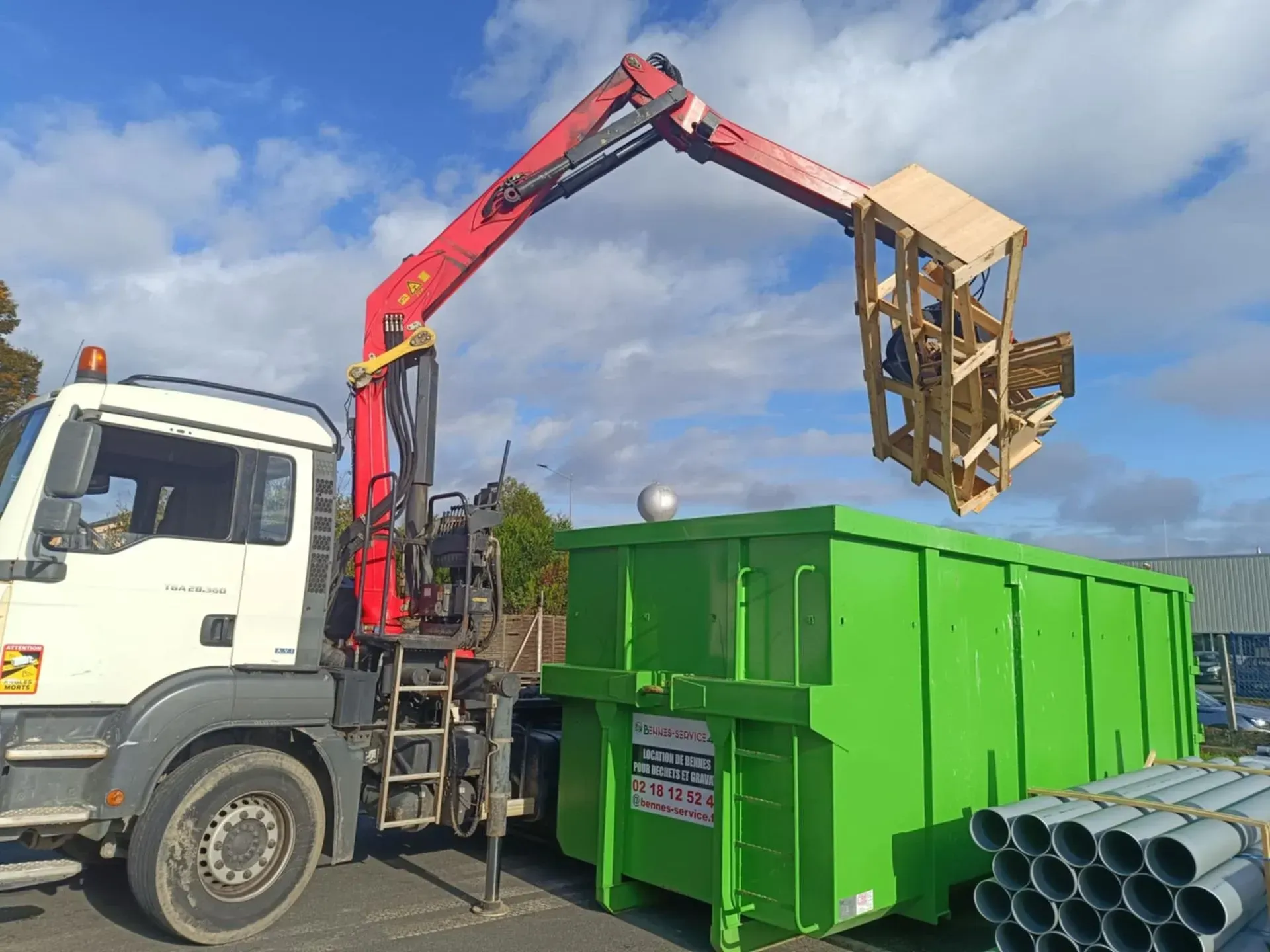 Un camion-grue soulève une structure en bois au-dessus d'une benne à ordures verte. Journée ensoleillée.