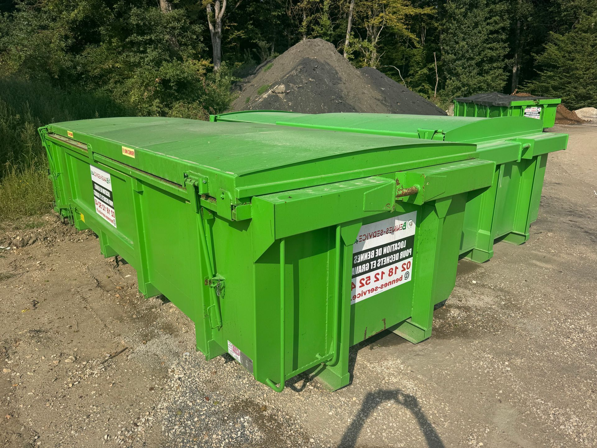 Deux bennes à ordures métalliques vert vif sont posées sur du gravier, avec un tas de matériaux sombres en arrière-plan.