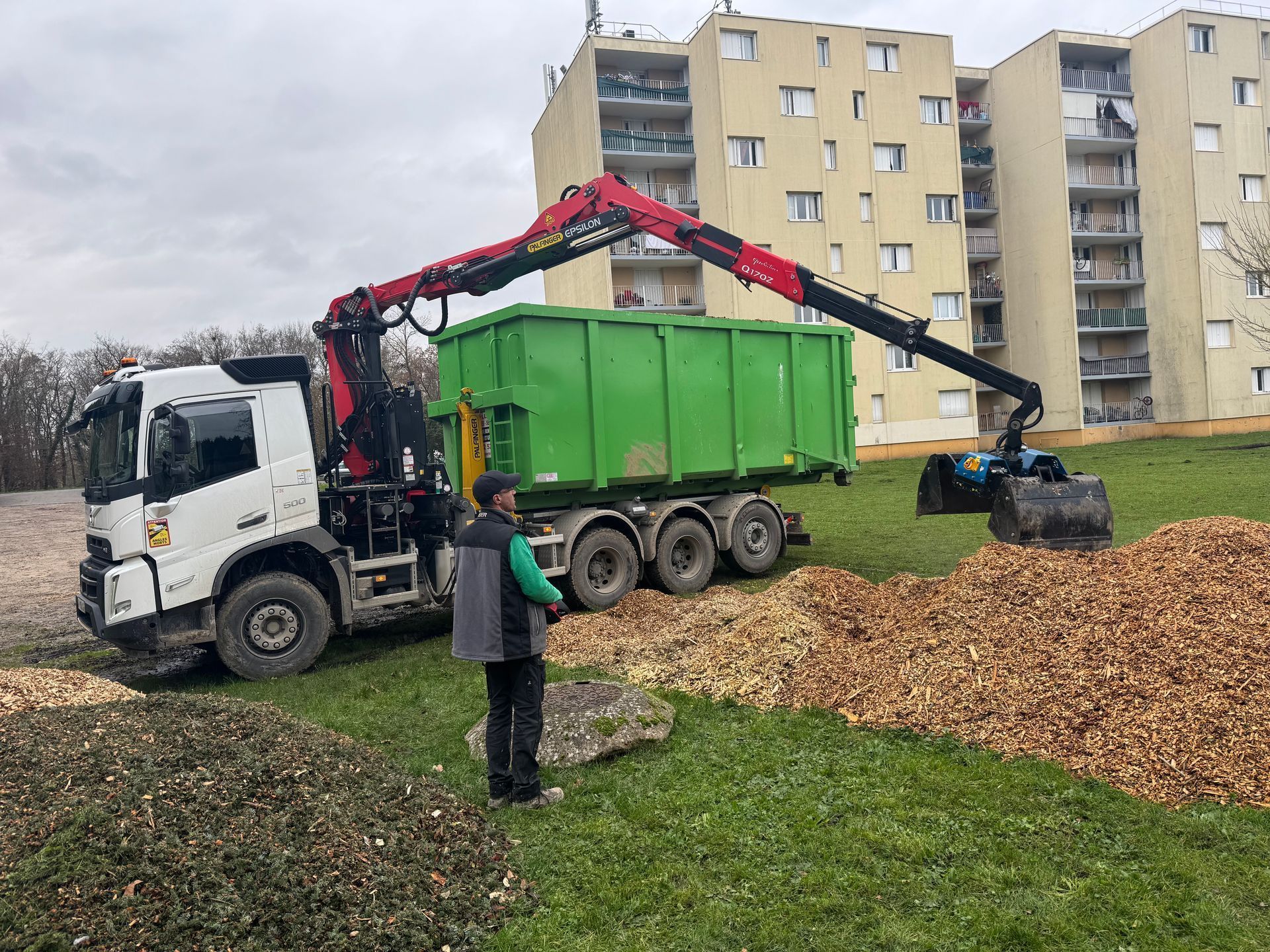 Un camion-grue charge des copeaux de bois dans une benne verte ; une personne observe la scène ; une zone herbeuse ; un bâtiment apparaît en arrière-plan.