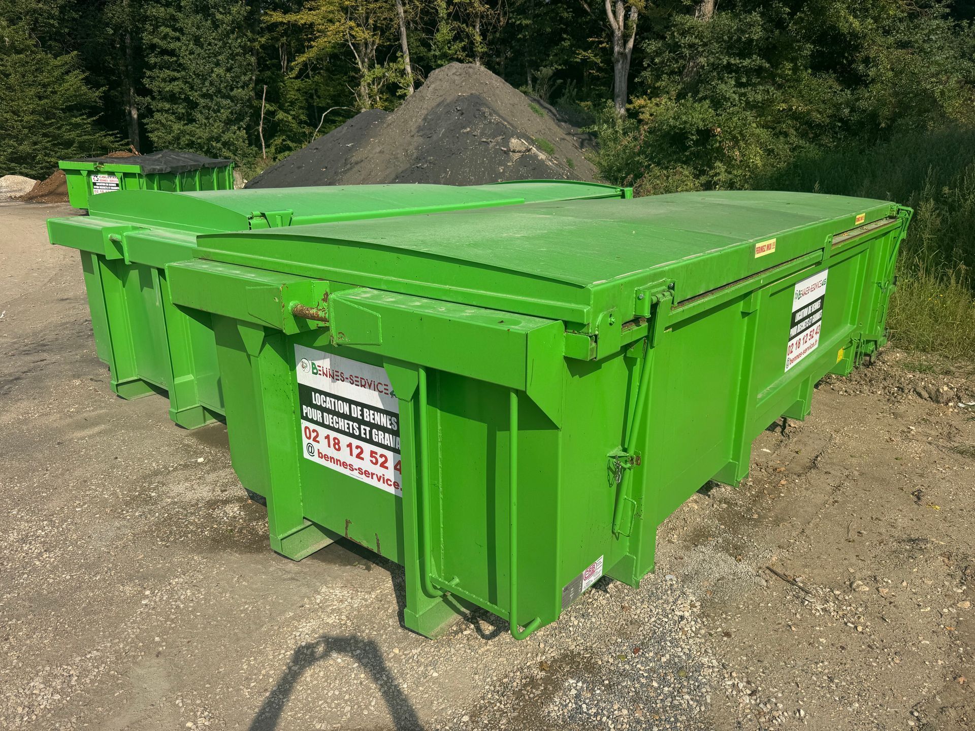 Conteneurs à déchets verts sur du gravier, avec un tas de matériaux sombres en arrière-plan.