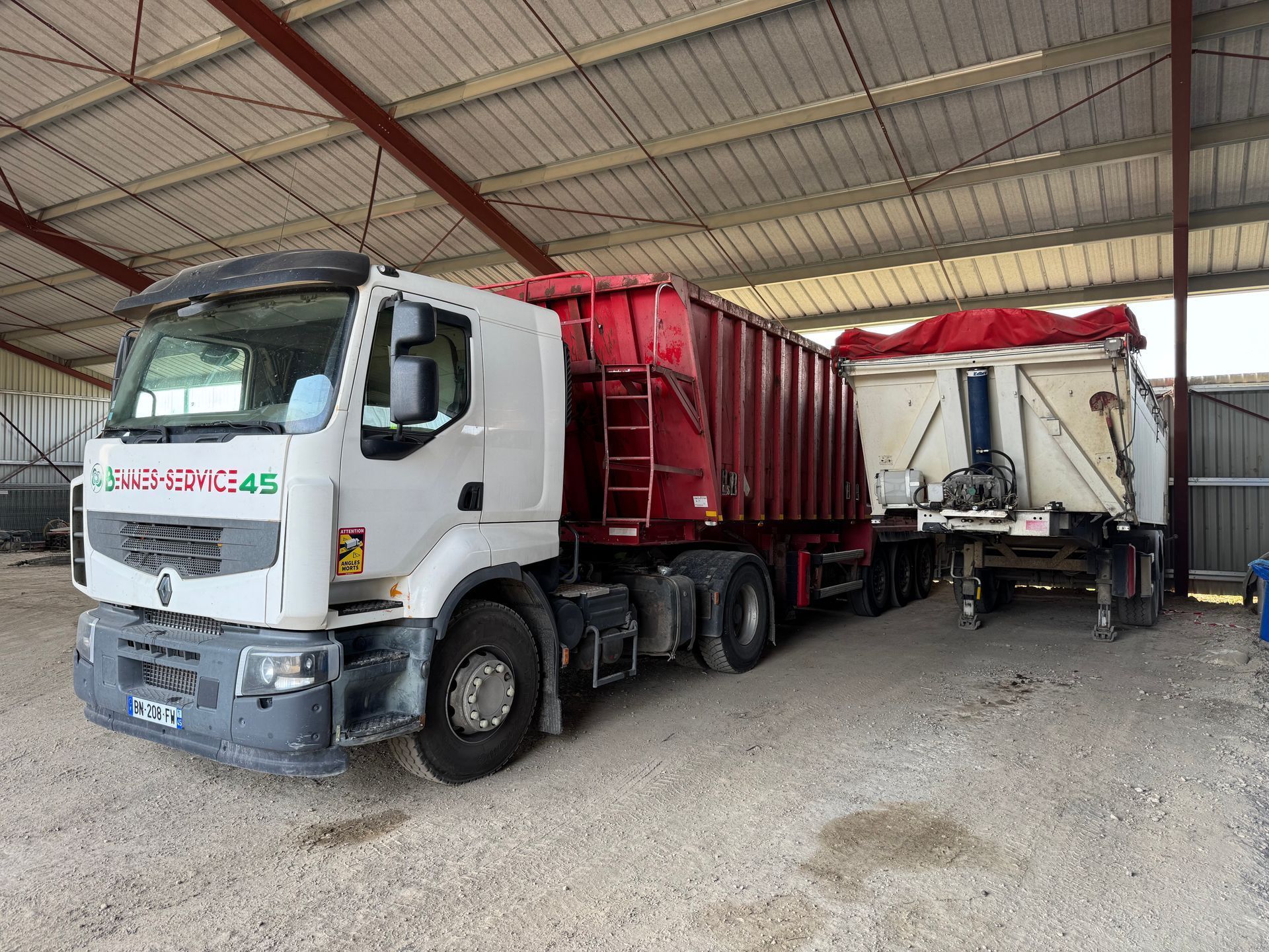 Un camion blanc avec un conteneur rouge et une remorque dans un hangar au sol de gravier.