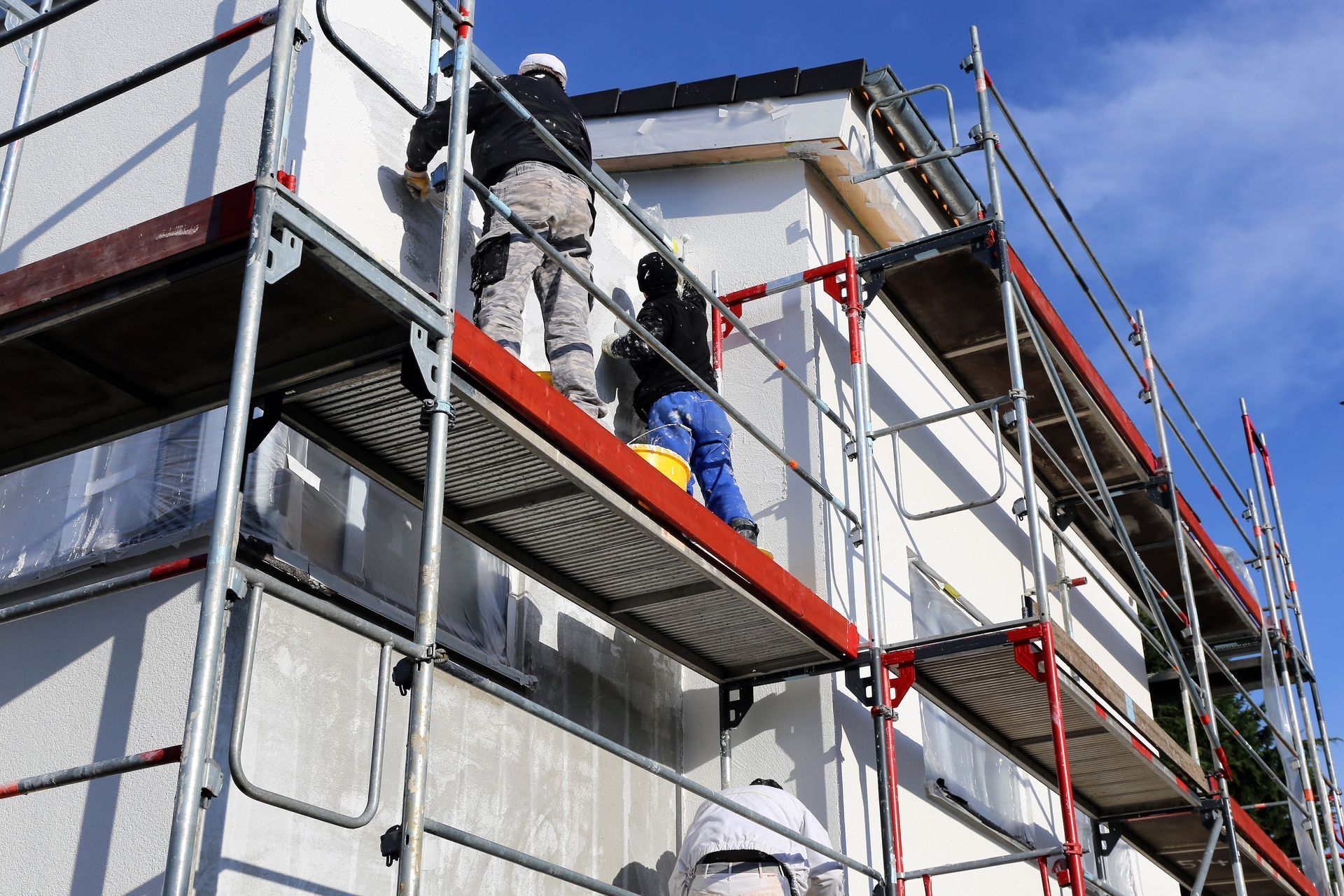 Bauarbeiter auf einem Gerüst, die unter blauem Himmel Material auf ein weisses Gebäude aufbringen.