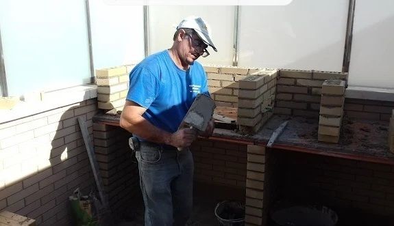 Hombre con camisa azul y sombrero, colocando ladrillos para construir una estructura, con herramientas y materiales cerca.